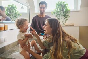 Lachende Mutter cremt ihr Kleinkind im Badezimmer ein, während der Vater im Hintergrund zufrieden zuschaut.