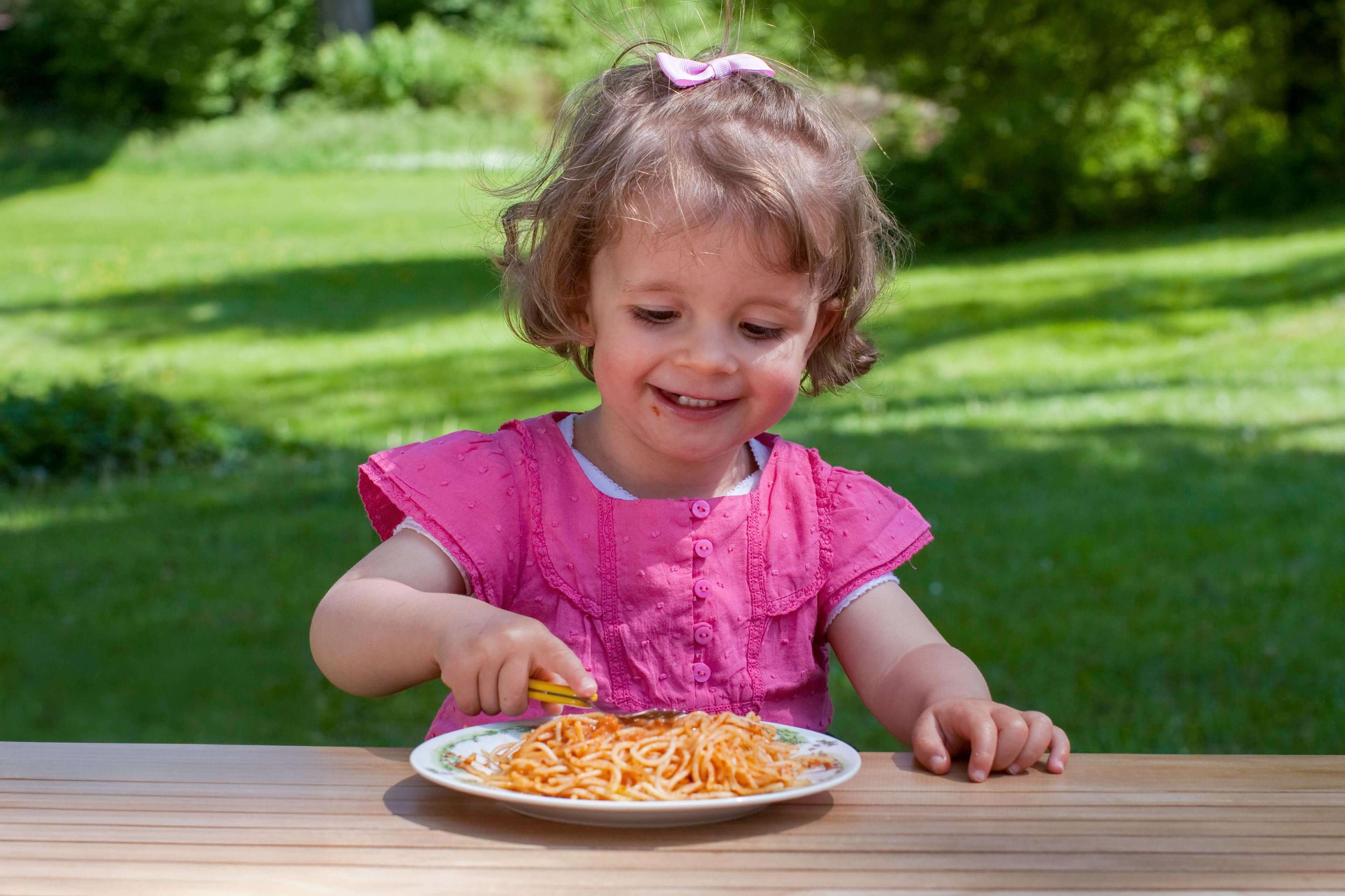Kind mit rosa Oberteil sitzt an Holztisch im Freien und isst Spaghetti von weißem Teller