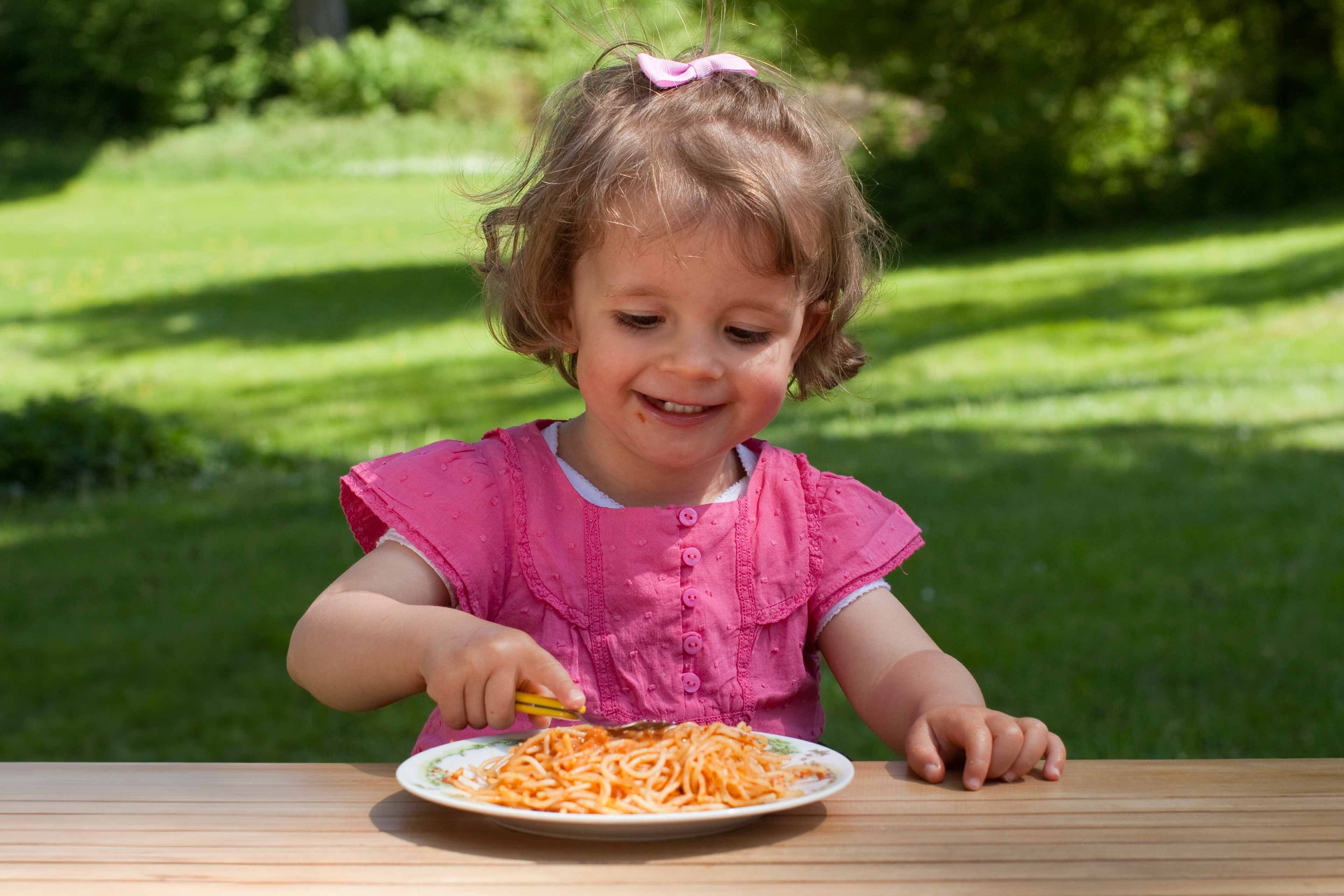 Ein kleines Kind in einem rosafarbenen Outfit sitzt an einem Tisch im Freien und isst Spaghetti.