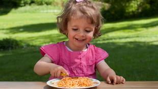 Ein kleines Kind in einem rosafarbenen Outfit sitzt an einem Tisch im Freien und isst Spaghetti.