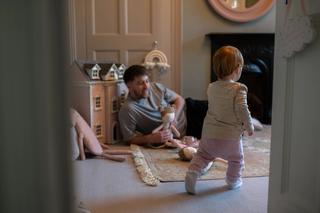 Father laying on the floor holding a doll and playing with toddler.