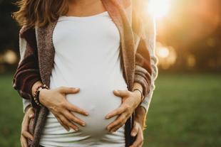 Die Hände einer Person, die sanft einen schwangeren Bauch streichelt, vor dem Hintergrund einer sonnenbeschienenen Freiluftkulisse.