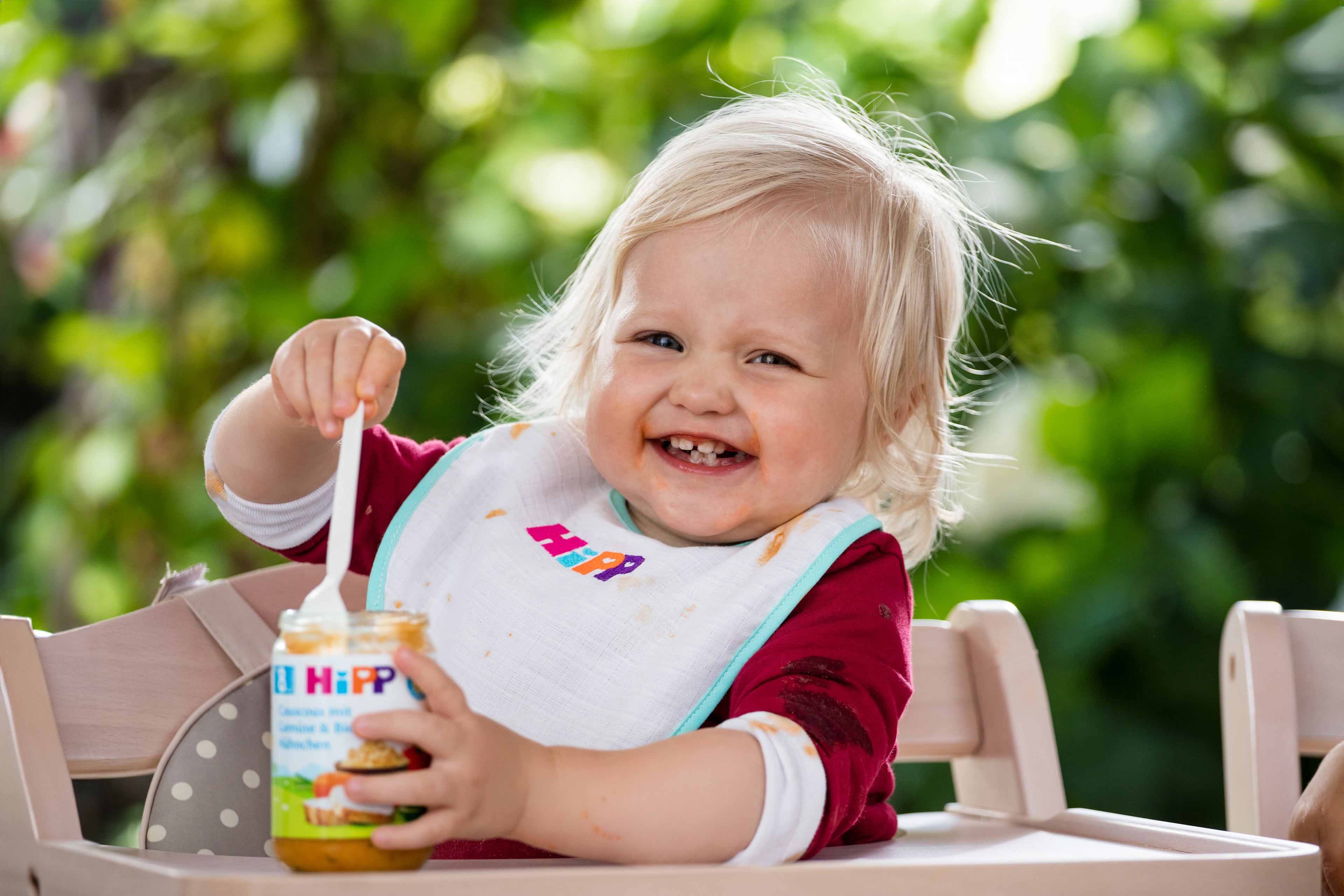 Ein fröhliches Kleinkind mit blondem Haar, das ein Lätzchen mit bunten Motiven trägt und ein Glas mit Essen mit der Aufschrift "Hipp" hält.