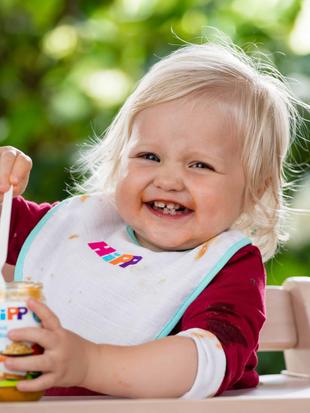 Ein fröhliches Kleinkind mit blondem Haar, das ein Lätzchen mit bunten Motiven trägt und ein Glas mit Essen mit der Aufschrift "Hipp" hält.