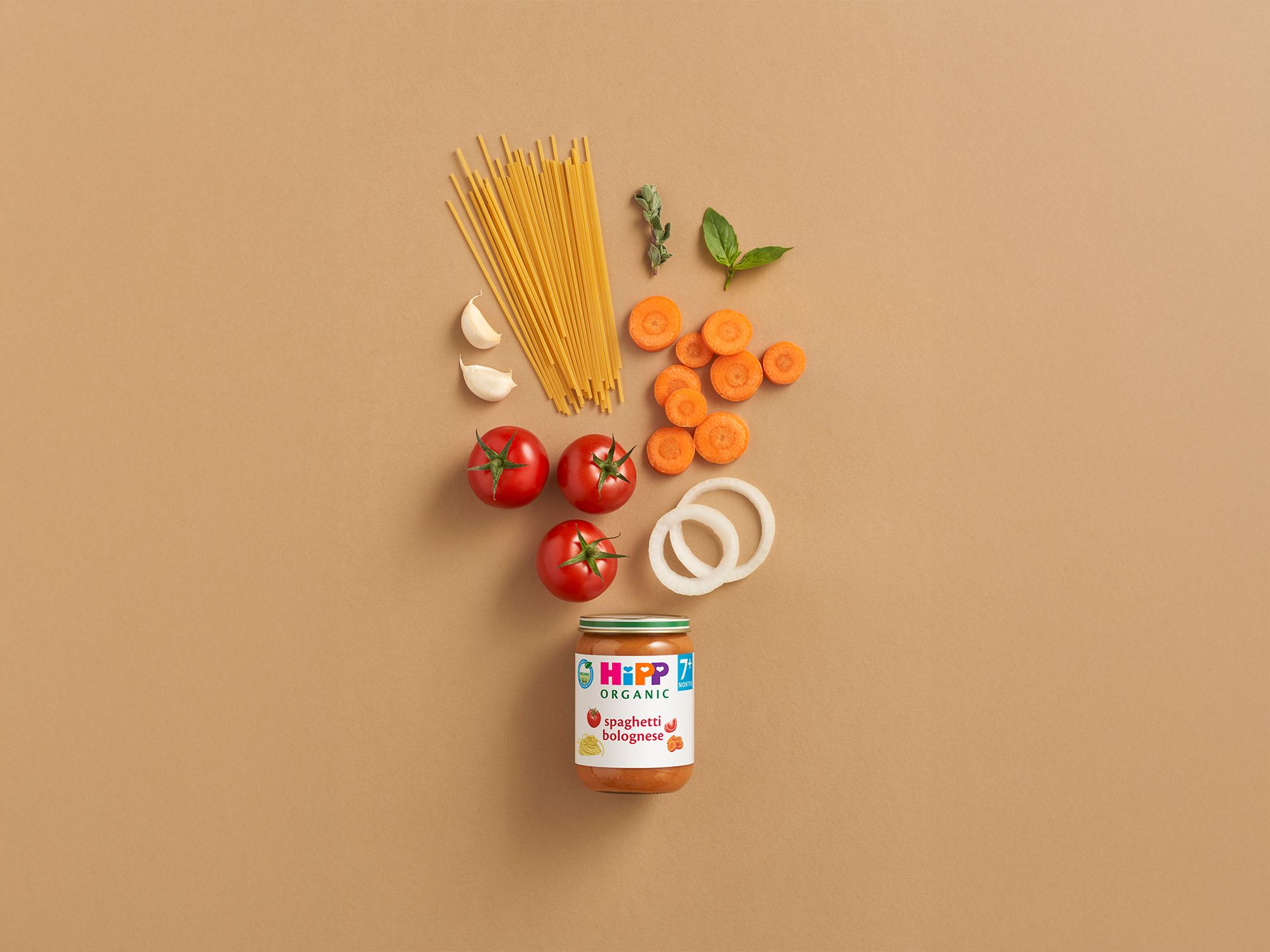 HiPP Organic Spaghetti Bolognese jar surrounded by tomatoes, carrots, spaghetti, onion, garlic and herbs on a neutral background
