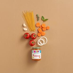 HiPP Organic Spaghetti Bolognese jar surrounded by tomatoes, carrots, spaghetti, onion, garlic and herbs on a neutral background