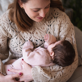 A mother wearing a cozy beige knit sweater gently cradles her baby, who is dressed in a soft pink outfit. The baby rests peacefully in her arms, sucking on their tiny hand, while the mother gazes down with a tender smile. The warm and intimate moment is captured in a softly lit setting with a cozy blanket and greenery in the background.
