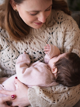 A mother wearing a cozy beige knit sweater gently cradles her baby, who is dressed in a soft pink outfit. The baby rests peacefully in her arms, sucking on their tiny hand, while the mother gazes down with a tender smile. The warm and intimate moment is captured in a softly lit setting with a cozy blanket and greenery in the background.
