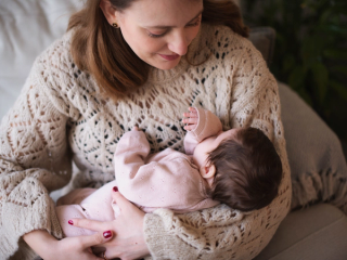 A mother wearing a cozy beige knit sweater gently cradles her baby, who is dressed in a soft pink outfit. The baby rests peacefully in her arms, sucking on their tiny hand, while the mother gazes down with a tender smile. The warm and intimate moment is captured in a softly lit setting with a cozy blanket and greenery in the background.