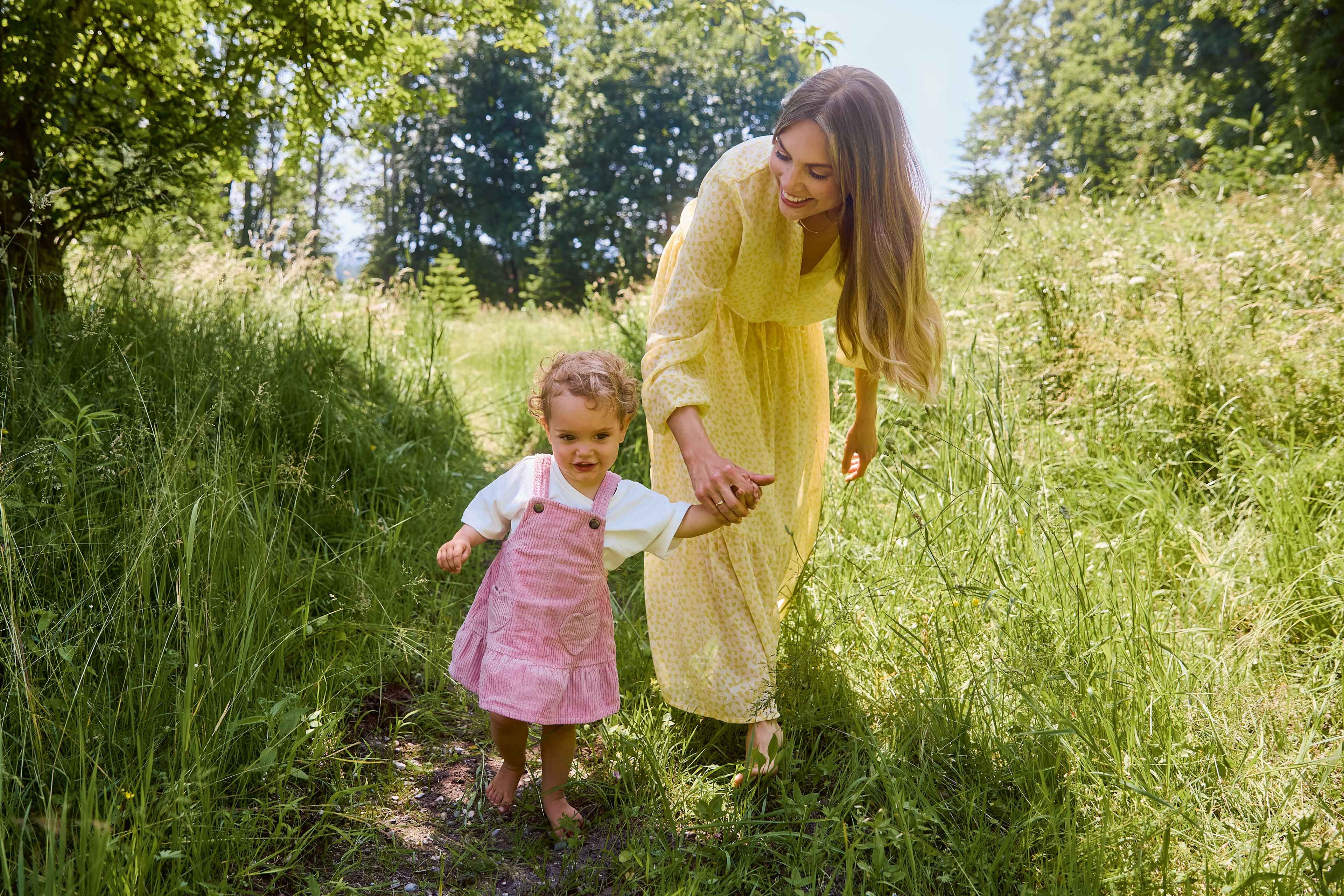Frau im gelben Kleid hält die Hand eines Kleinkinds im rosa Latzkleid beim Spaziergang auf einem grasbewachsenen Pfad