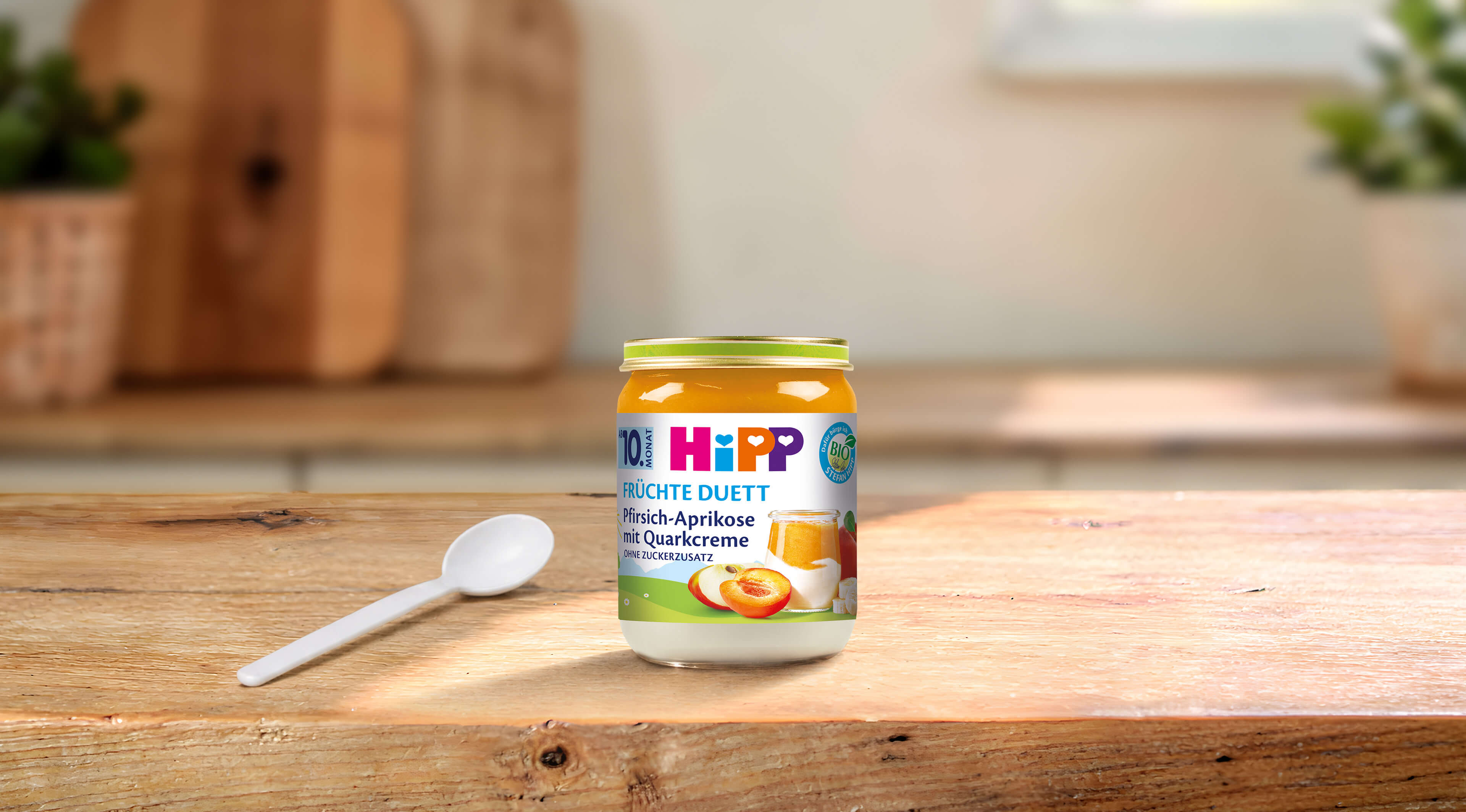HiPP Früchte-Duett Bio-Gläschen Pfirsich-Aprikose mit Quarkcreme ab 10 Monaten auf Holztisch mit Löffel in heller Küche