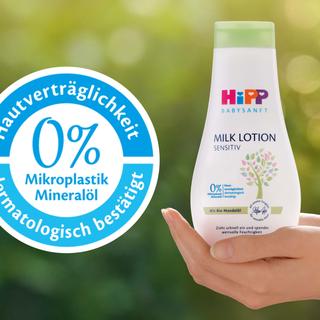 Eine Flasche eines Babypflegeprodukts mit der Aufschrift "Hipp Baby Sensitive" und einem Etikett mit der Aufschrift "0% Mikroplastik Mineralol" sowie eine Hand, die das Produkt vor einem grünen Hintergrund hält.