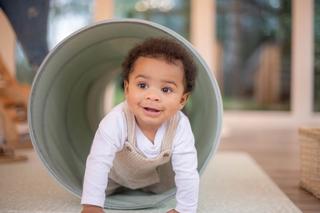 A smiling baby crawls through a soft play tunnel, wearing a white long-sleeve shirt and beige corduroy overalls. The setting appears to be a bright indoor play area with large windows and cozy decor