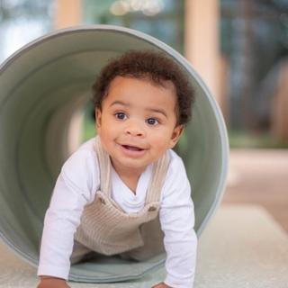A smiling baby crawls through a soft play tunnel, wearing a white long-sleeve shirt and beige corduroy overalls. The setting appears to be a bright indoor play area with large windows and cozy decor.
