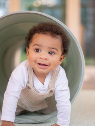 A smiling baby crawls through a soft play tunnel, wearing a white long-sleeve shirt and beige corduroy overalls. The setting appears to be a bright indoor play area with large windows and cozy decor.