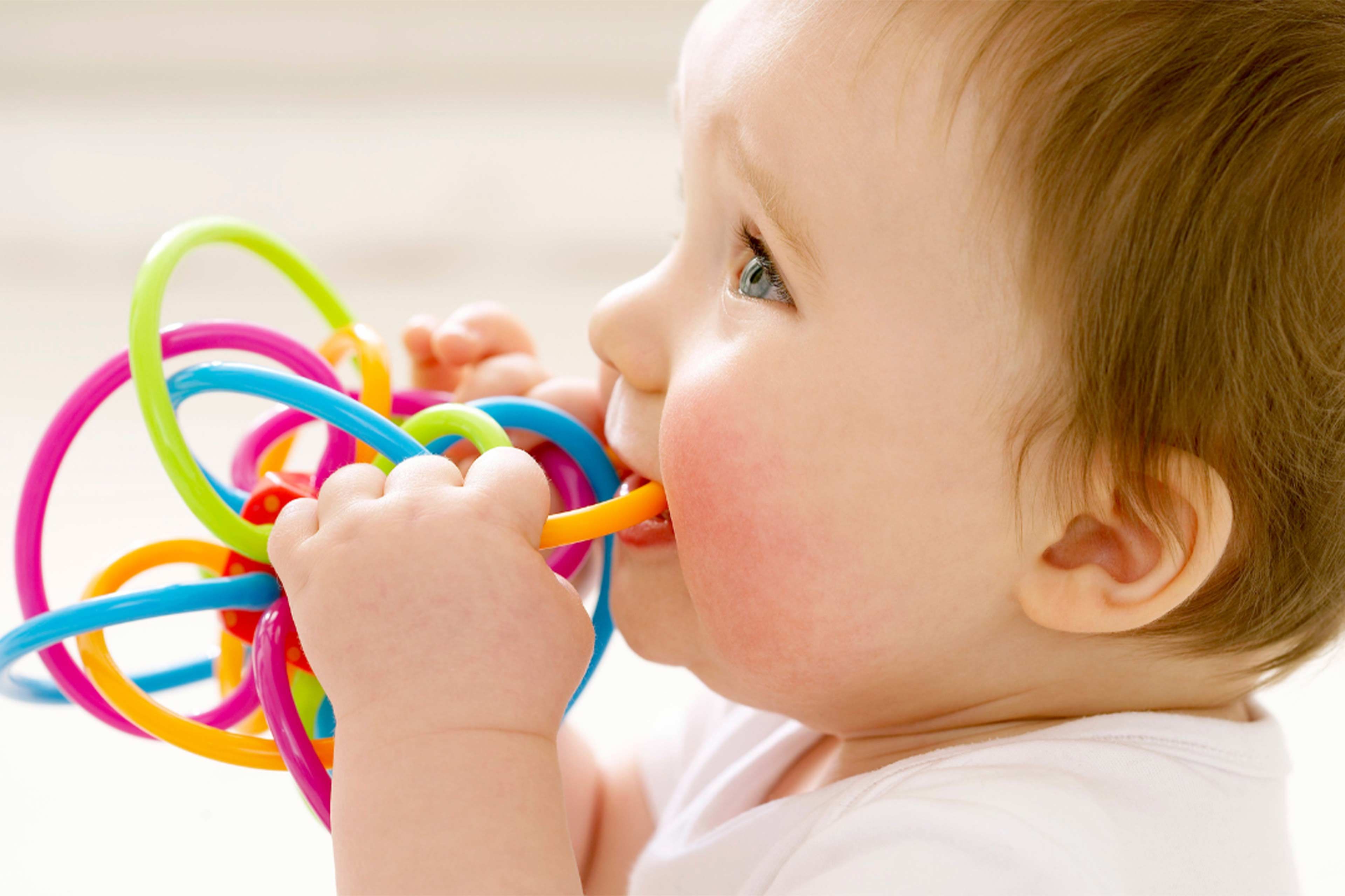 Lächelndes Baby knabbert an einem bunten Beißspielzeug zur Zahnungshilfe vor hellem, weichem Hintergrund