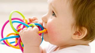 Lächelndes Baby knabbert an einem bunten Beißspielzeug zur Zahnungshilfe vor hellem, weichem Hintergrund