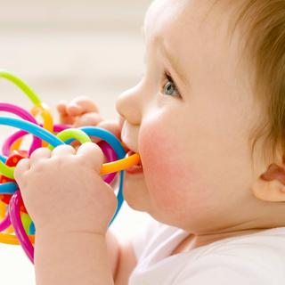 Lächelndes Baby knabbert an einem bunten Beißspielzeug zur Zahnungshilfe vor hellem, weichem Hintergrund