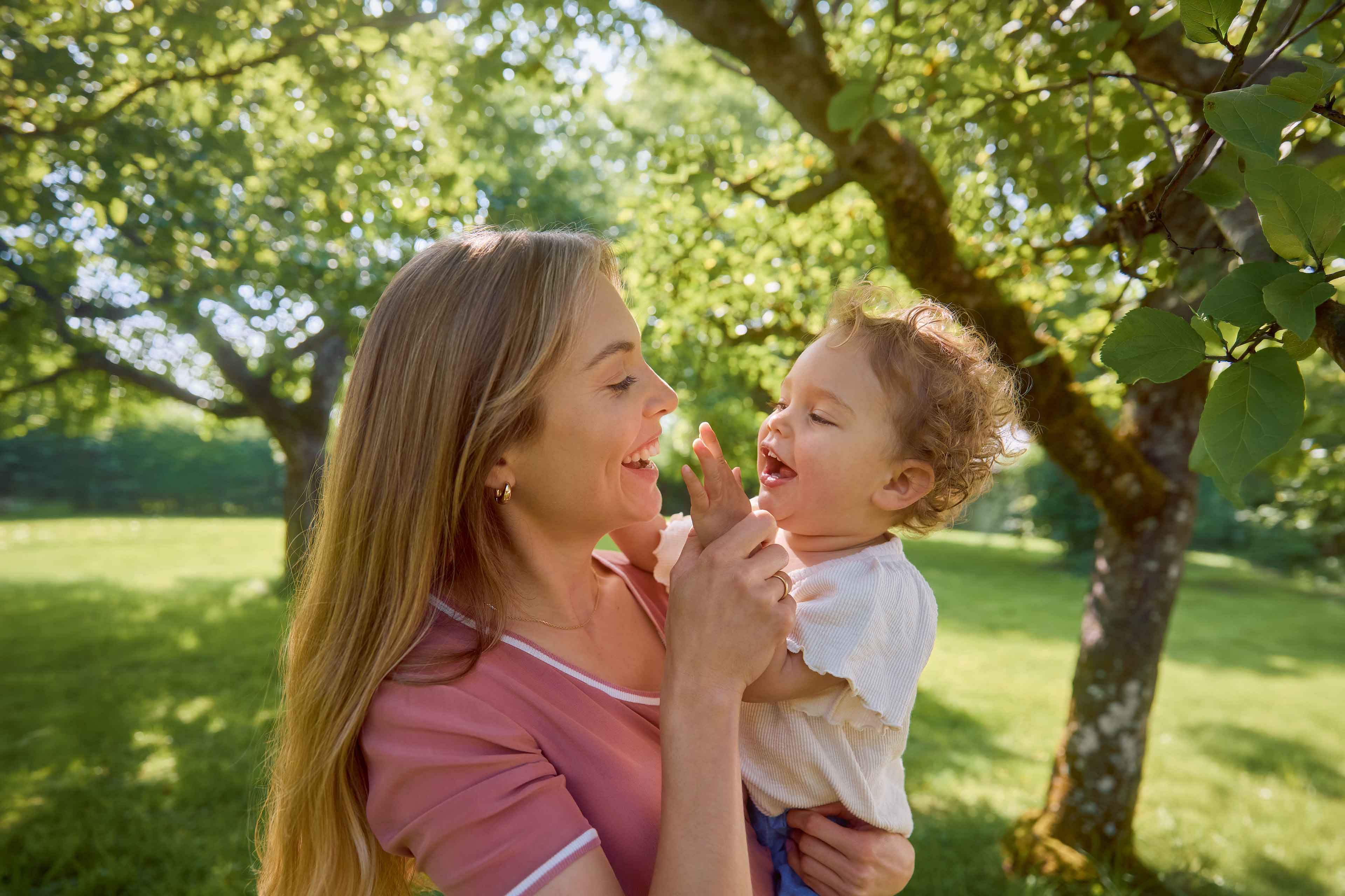 Lachende Mutter hält ihr Baby im grünen Park im Arm, beide schauen sich liebevoll an und genießen die gemeinsame Zeit.