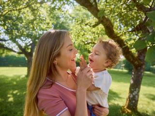 Lachende Mutter hält ihr Baby im grünen Park im Arm, beide schauen sich liebevoll an und genießen die gemeinsame Zeit.