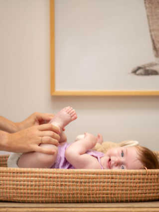 Mum changing baby in purple dress nappy, baby is smiling whilst holding her feet