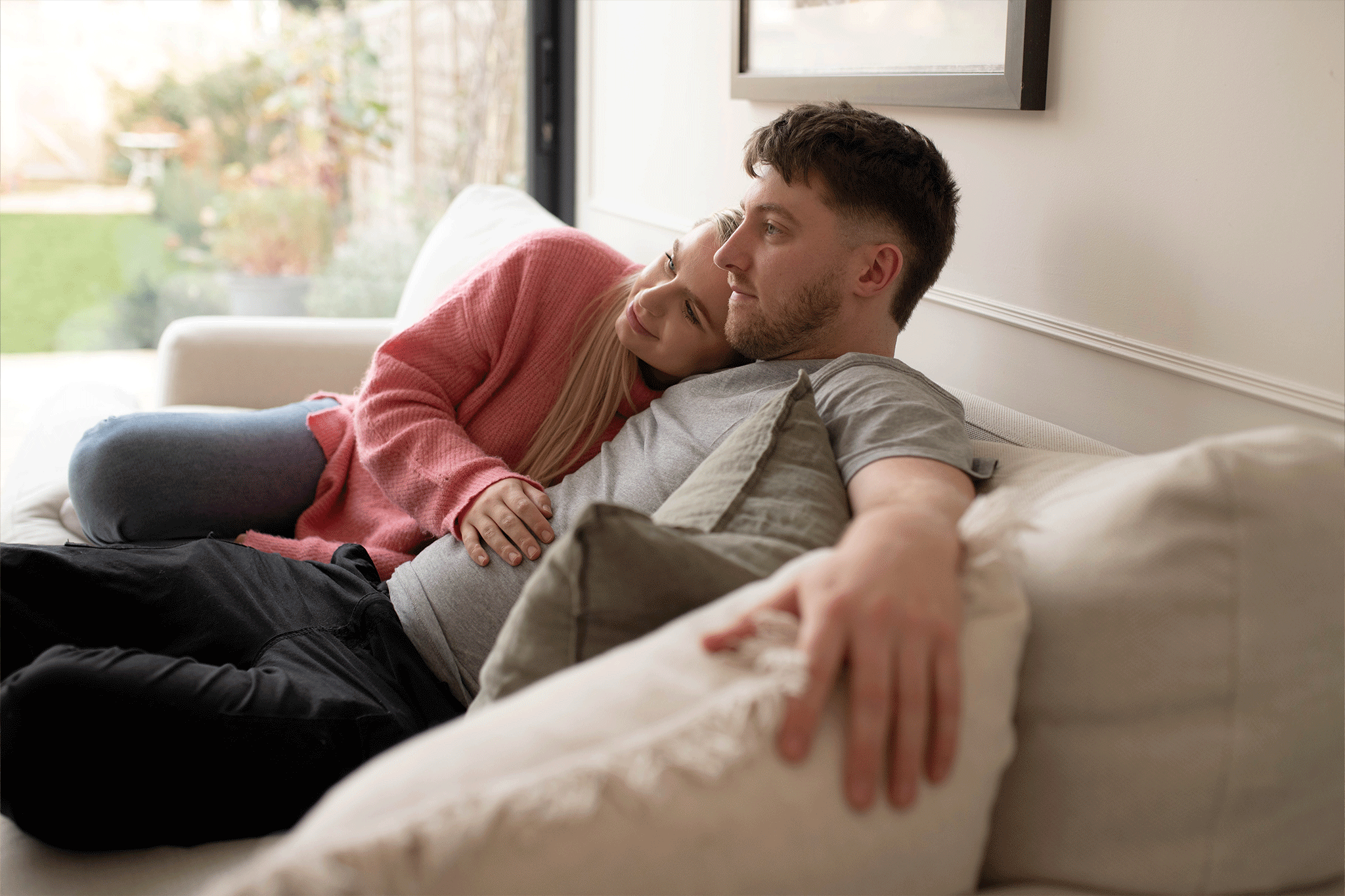 A couple relaxing together on a light beige sofa indoors. A woman wearing a pink knit cardigan rests her head affectionately on a man's shoulder while placing her hand on his stomach. The man, wearing a grey t-shirt and black pants, leans back with his arm resting along the cushions while gazing thoughtfully into the room. Light streams in from large glass doors in the background.