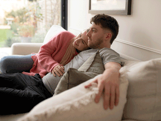 A couple relaxing together on a light beige sofa indoors. A woman wearing a pink knit cardigan rests her head affectionately on a man's shoulder while placing her hand on his stomach. The man, wearing a grey t-shirt and black pants, leans back with his arm resting along the cushions while gazing thoughtfully into the room. Light streams in from large glass doors in the background.
