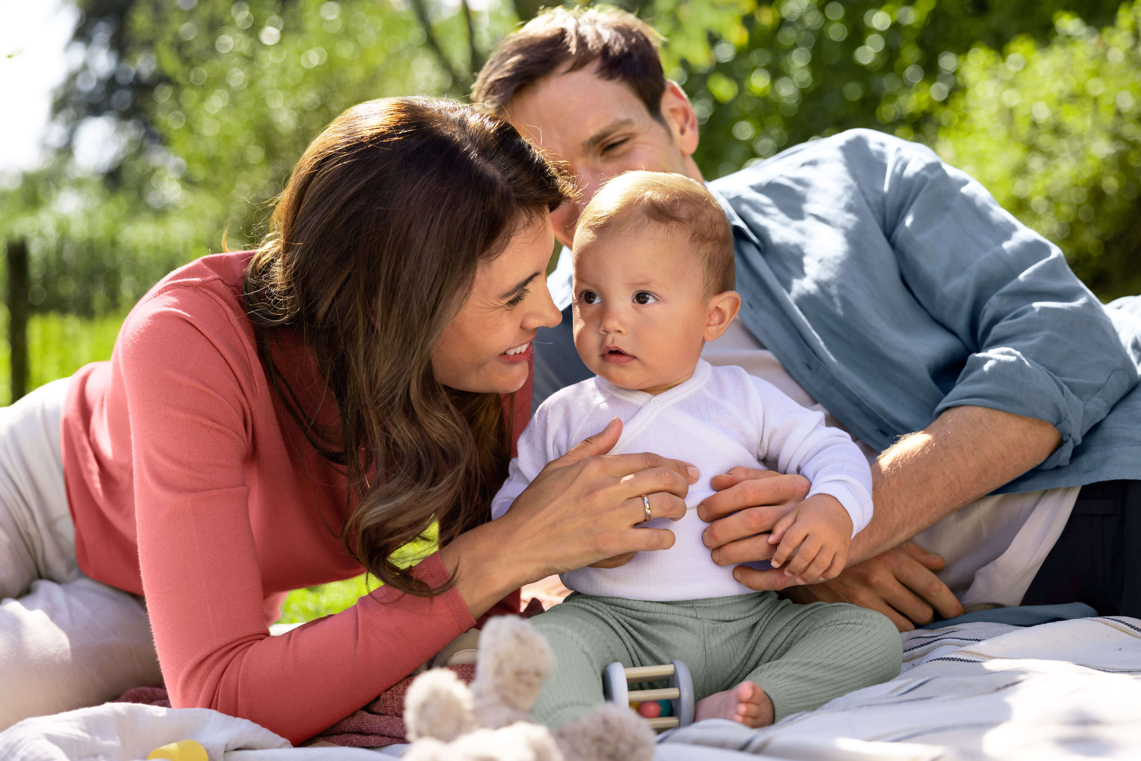 Eltern kuscheln mit ihrem sitzenden Baby auf einer Decke im Grünen – entspanntes Familienpicknick für gesundes Aufwachsen.