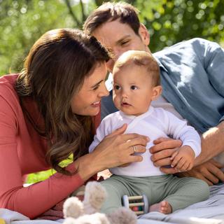 Eltern kuscheln mit ihrem sitzenden Baby auf einer Decke im Grünen – entspanntes Familienpicknick für gesundes Aufwachsen.