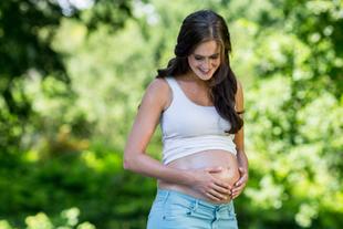 Eine Frau in einer natürlichen Umgebung, die sanft ihren Bauch berührt und in freudiger Stimmung zu sein scheint.