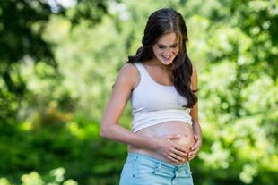 Eine Frau in einer natürlichen Umgebung, die sanft ihren Bauch berührt und in freudiger Stimmung zu sein scheint.