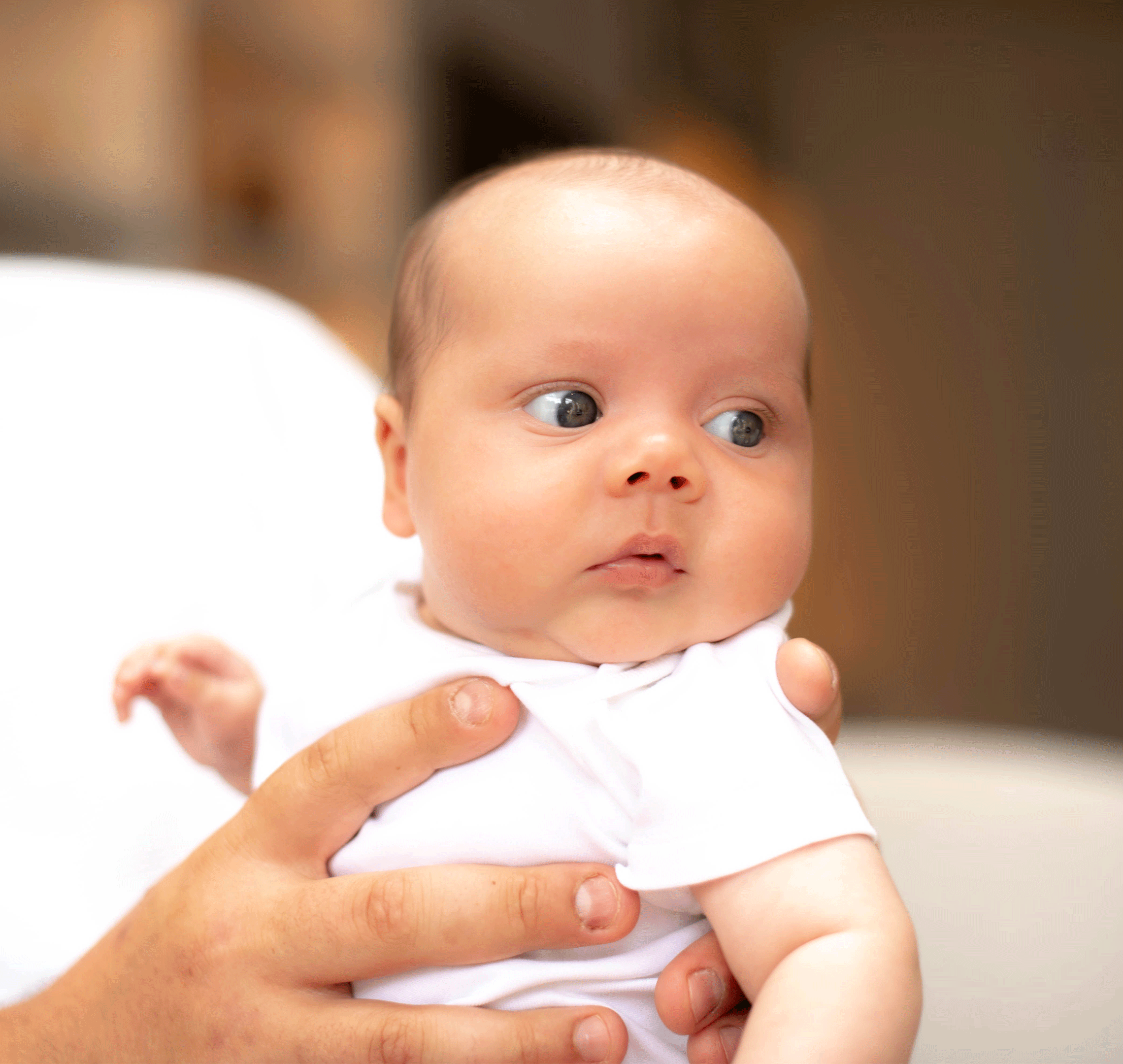 A close-up photograph of a young baby wearing a white vest, being cradled in an adult's hands. The baby is looking off to the right with wide, dark eyes, showing the top and side of their head clearly.