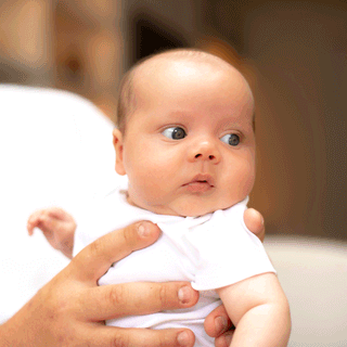 A close-up photograph of a young baby wearing a white vest, being cradled in an adult's hands. The baby is looking off to the right with wide, dark eyes, showing the top and side of their head clearly.
