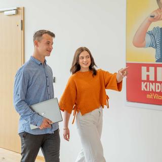 Zwei junge Erwachsene gehen im Flur, die Frau zeigt auf ein Plakat an der Wand