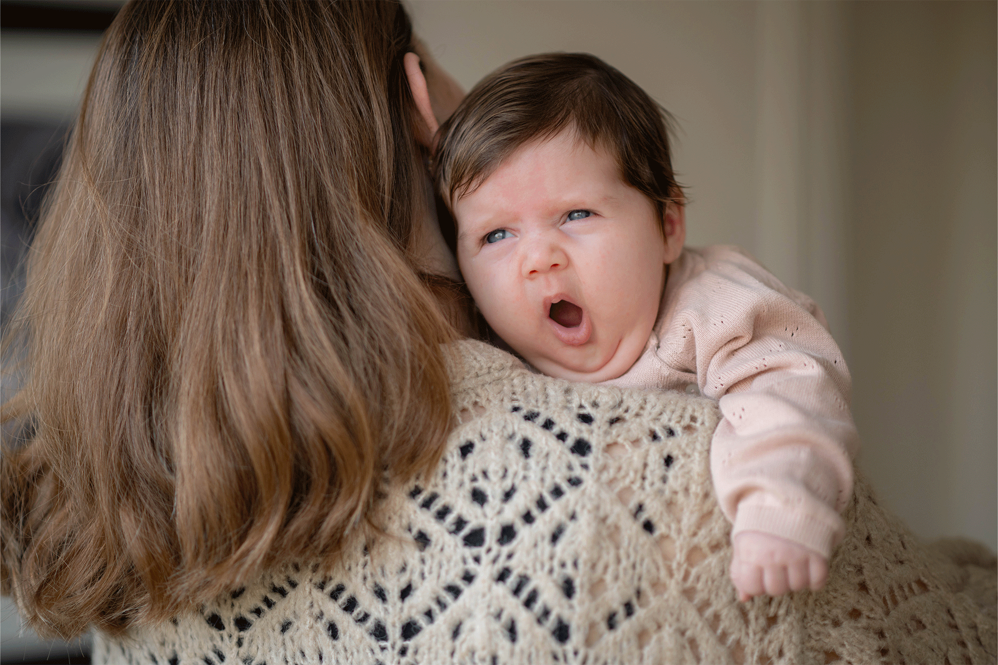 A close-up shot of a newborn baby with dark hair and blue eyes resting over an adult's shoulder, mid-yawn with an open mouth. The baby is wearing a light pink long-sleeved sweater, and the adult, seen from behind, wears a beige patterned knit sweater.