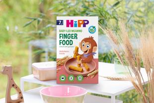 Packung Hipp Baby-led Weaning Finger Food mit Illustration eines Affen, der Sticks hält, auf einem weißen Tisch mit Schalen und Pflanzen im Hintergrund