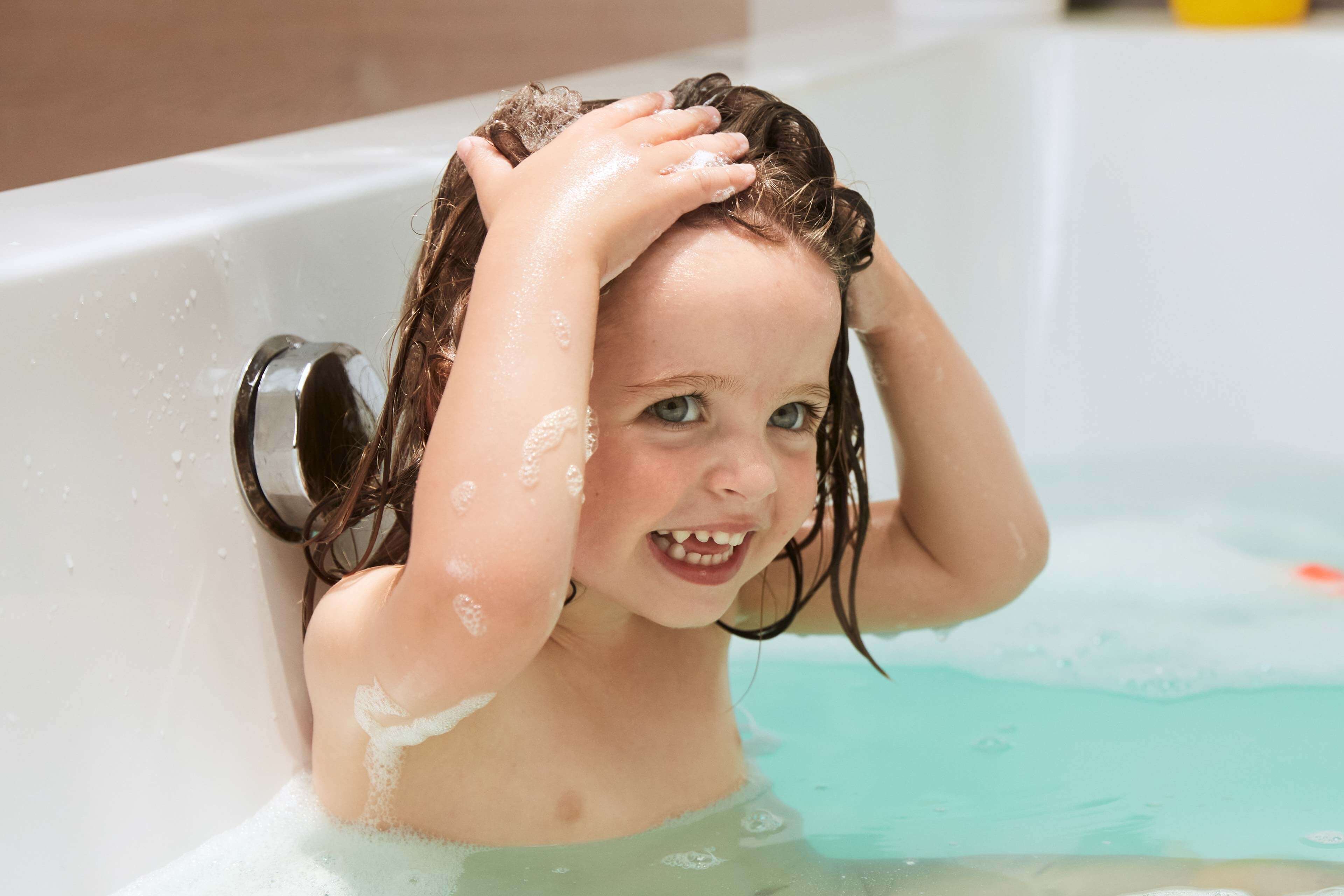 Ein kleines Kind in einer Badewanne, das mit Seifenblasen spielt und sein Haar hält.