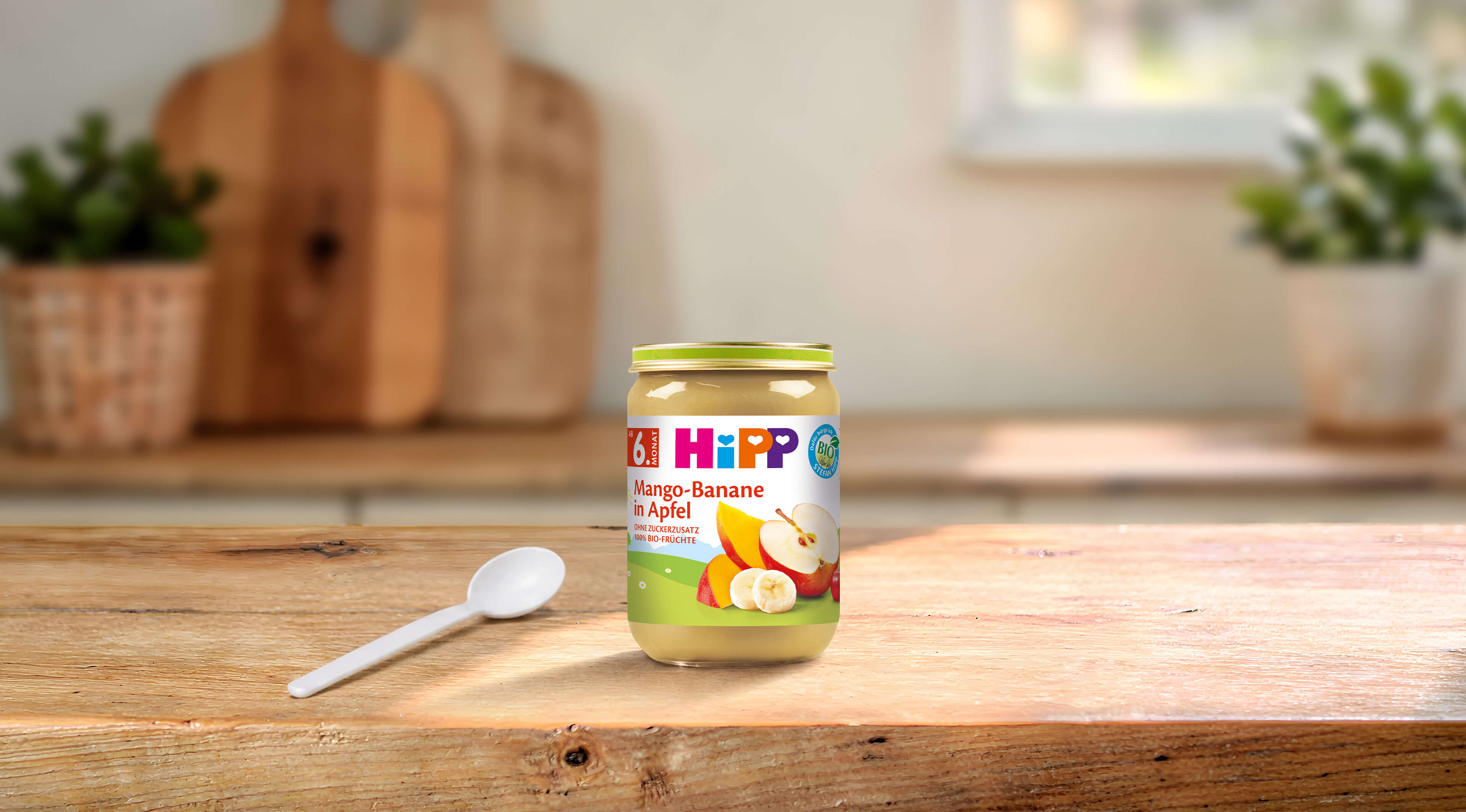 HiPP Bio-Gläschen Mango-Banane in Apfel ab 6. Monat auf Holztisch mit Löffel in heller Küche