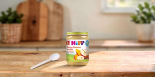 HiPP Bio-Gläschen Mango-Banane in Apfel ab 6. Monat auf Holztisch mit Löffel in heller Küche