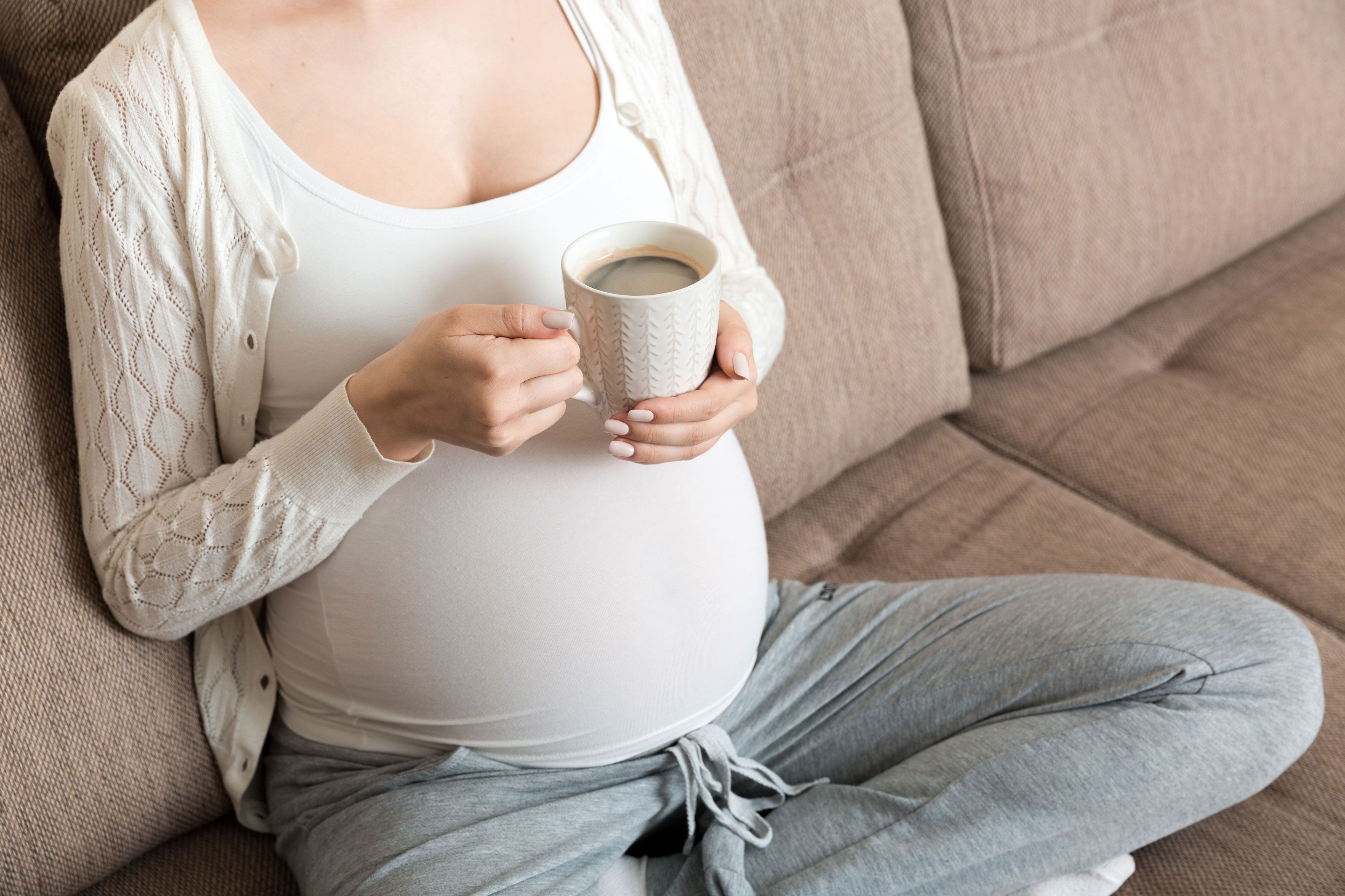 Eine Person hält einen weißen Becher mit einem dunklen Getränk, vermutlich Kaffee, vor einem neutralen Hintergrund.