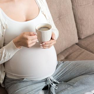 Eine Person hält einen weißen Becher mit einem dunklen Getränk, vermutlich Kaffee, vor einem neutralen Hintergrund.