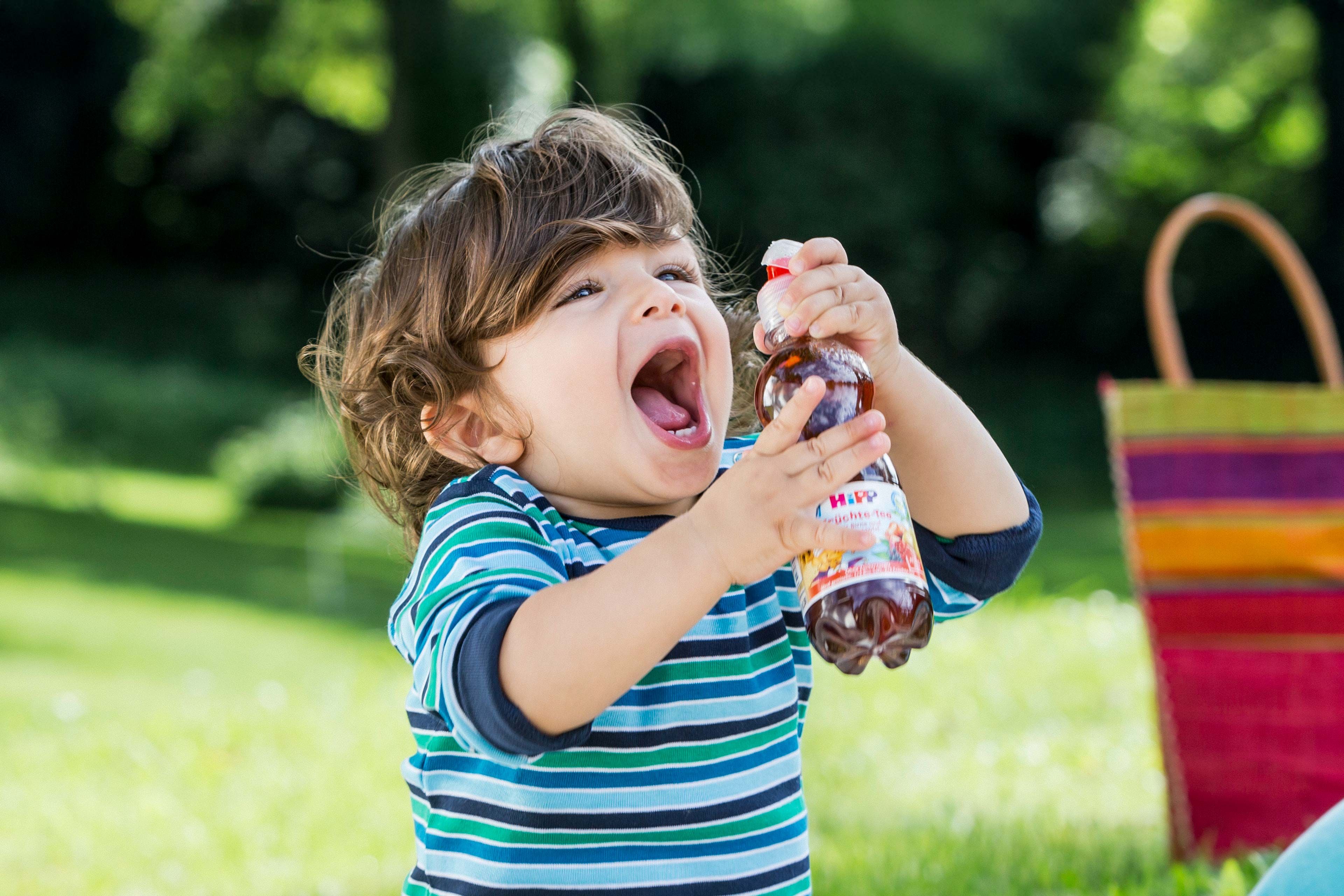 Lachendes Kleinkind im Grünen hält begeistert ein HiPP Bio-Kindergetränk in der Hand
