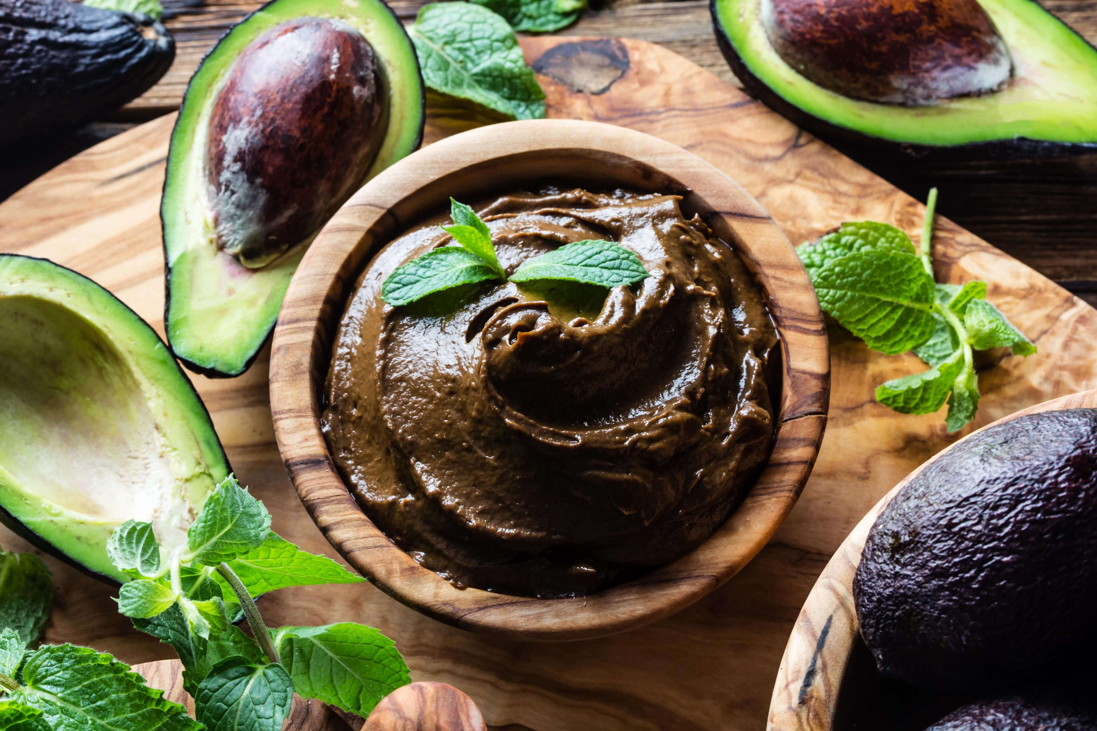 Eine Holzschale, gefüllt mit einer cremefarbenen Masse, garniert mit einem Minzblatt, umgeben von halbierten Avocados und frischen Minzblättern.