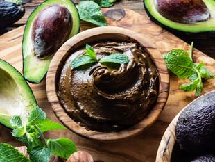 Eine Holzschale, gefüllt mit einer cremefarbenen Masse, garniert mit einem Minzblatt, umgeben von halbierten Avocados und frischen Minzblättern.
