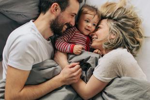 Mama und Papa kuscheln entspannt im Bett mit ihrem lachenden Baby und genießen Nähe in der Stillzeit