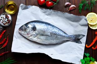 Ein frischer Fisch auf weißem Papier, umgeben von verschiedenen Zutaten wie Chilischoten, Zitrone, Knoblauch und Kräutern, alles vor einem Holzhintergrund.