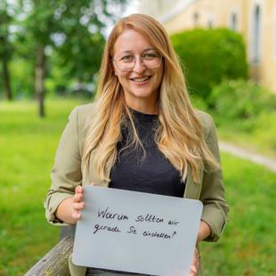 Eine HiPP Mitarbeiterin hält ein Schild mit der handgeschriebenen Frage 'Warum sollten wir gerade Sie einstellen' und lächelt in die Kamera