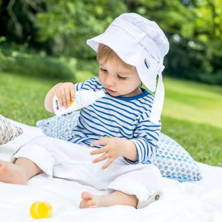 Ein kleines Kind mit weißem Hut und gestreiftem Hemd, das Sonnencreme aus einer weißen Flasche aufträgt.
