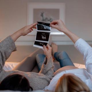 Two people lying on a bed, one visibly pregnant, holding up and looking at ultrasound images together in a cozy, softly lit room.