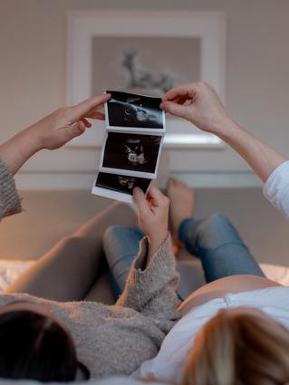 Two people lying on a bed, one visibly pregnant, holding up and looking at ultrasound images together in a cozy, softly lit room.